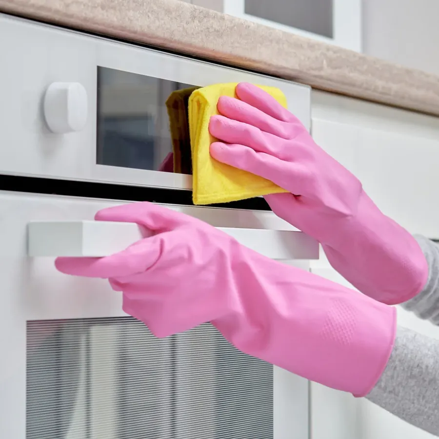 Pink gloved hands wiping a white oven door with a yellow cloth