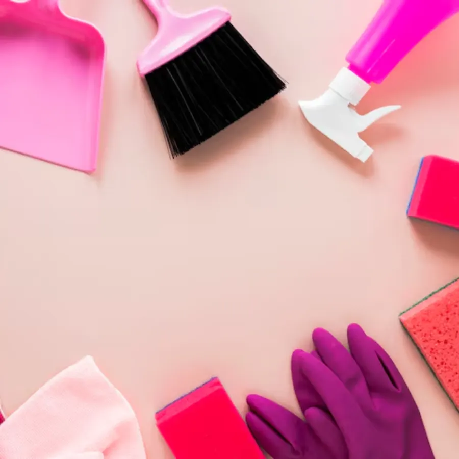Pink dustpan, hand brush, spray bottle, sponges, and gloves laid out on a pink background