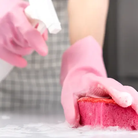 Gloved hand scrubbing a counter with a soapy pink sponge while holding a spray bottle