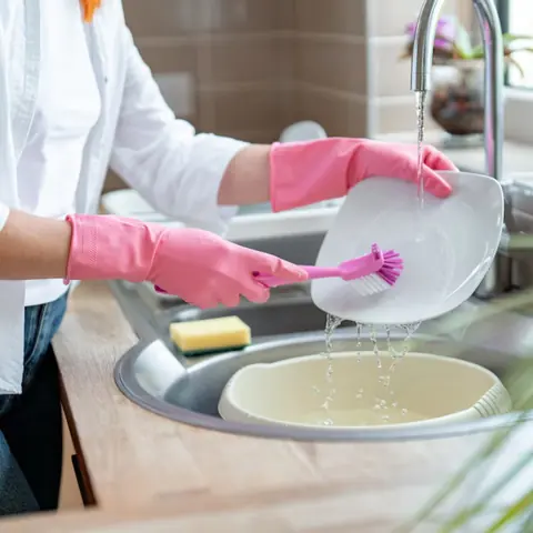 Pink gloved hands scrubbing a white plate with a dish brush at the kitchen sink