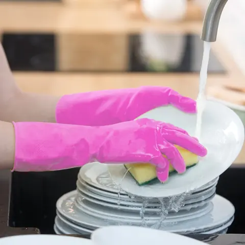 Pink gloved hands washing a stack of white plates under a kitchen faucet